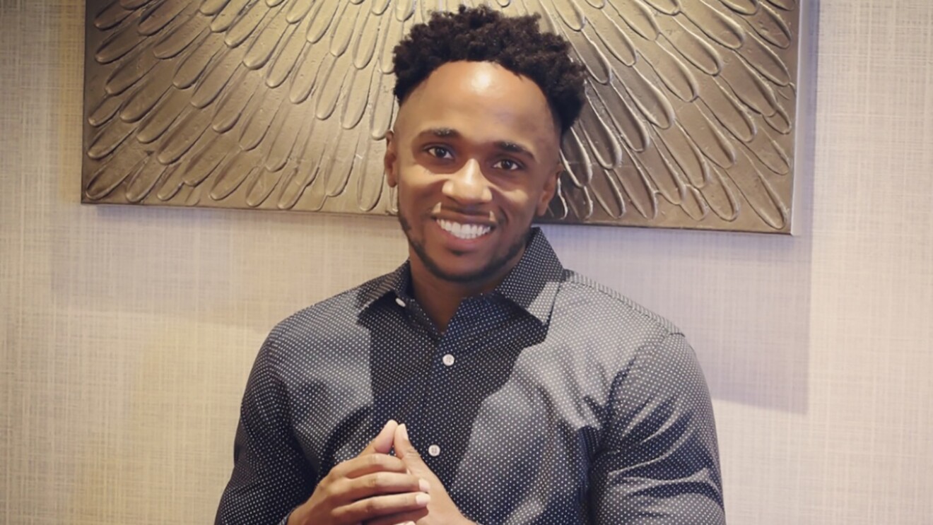 A Black man wearing a button up shirt smiles at the camera.