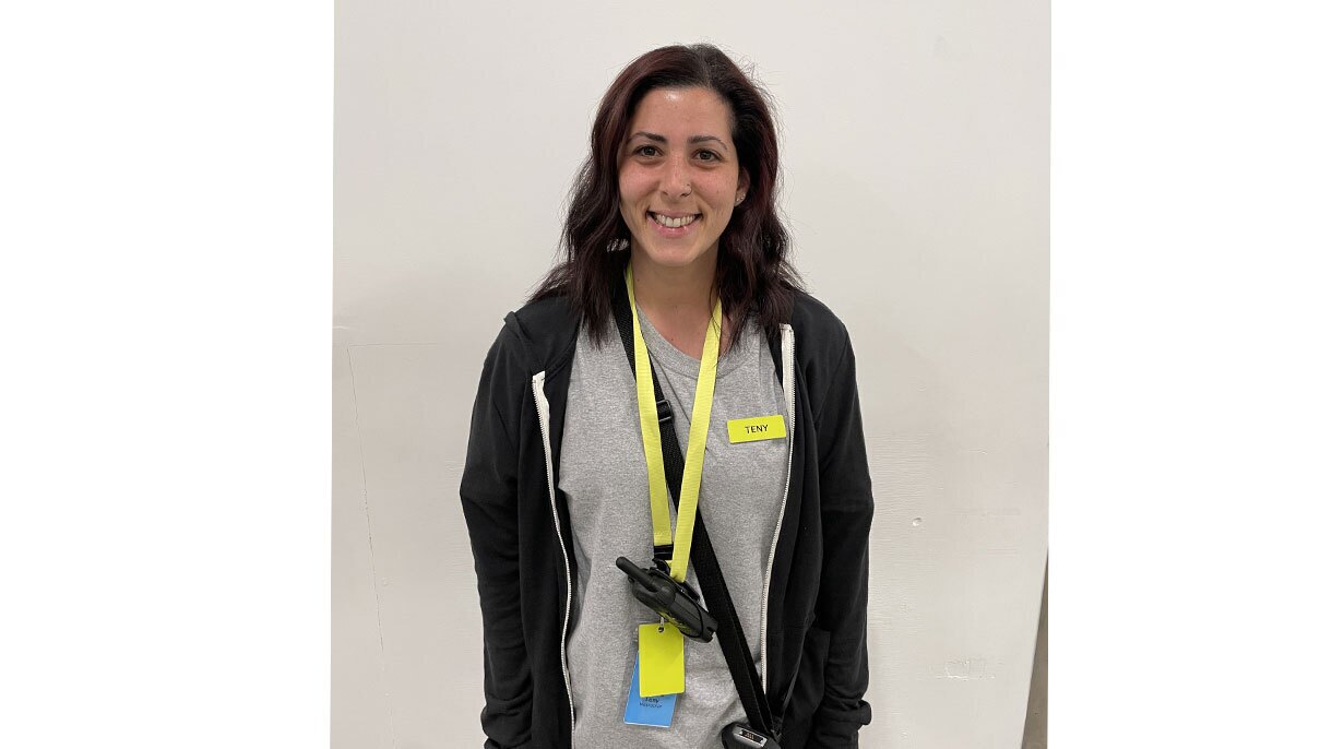 An image of a woman smiling for a photo standing in front of a white wall. She has a neon lanyard around her neck with a walkie talkie attached and a neon yellow name tag.