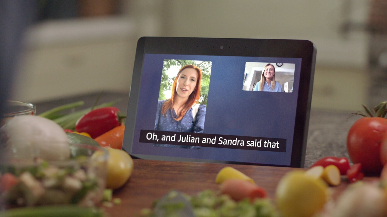 A video call featuring two women on an Amazon Echo Show 8 with call captioning sits atop a kitchen counter surrounded by vegetables.