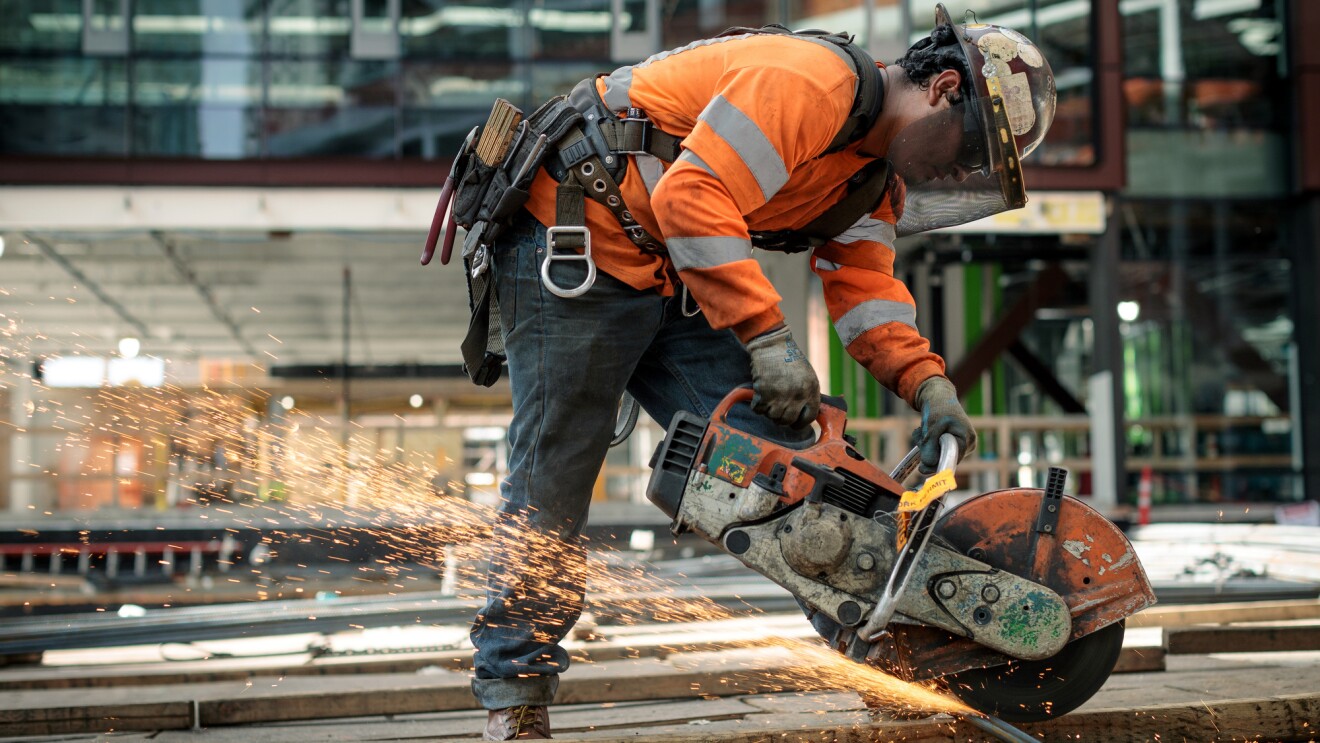 A man in a construction helmet and orange safety jacket is photographed at a construction site on Amazon's campus in Seattle, WA. He is using a circular saw to cut through metal. 