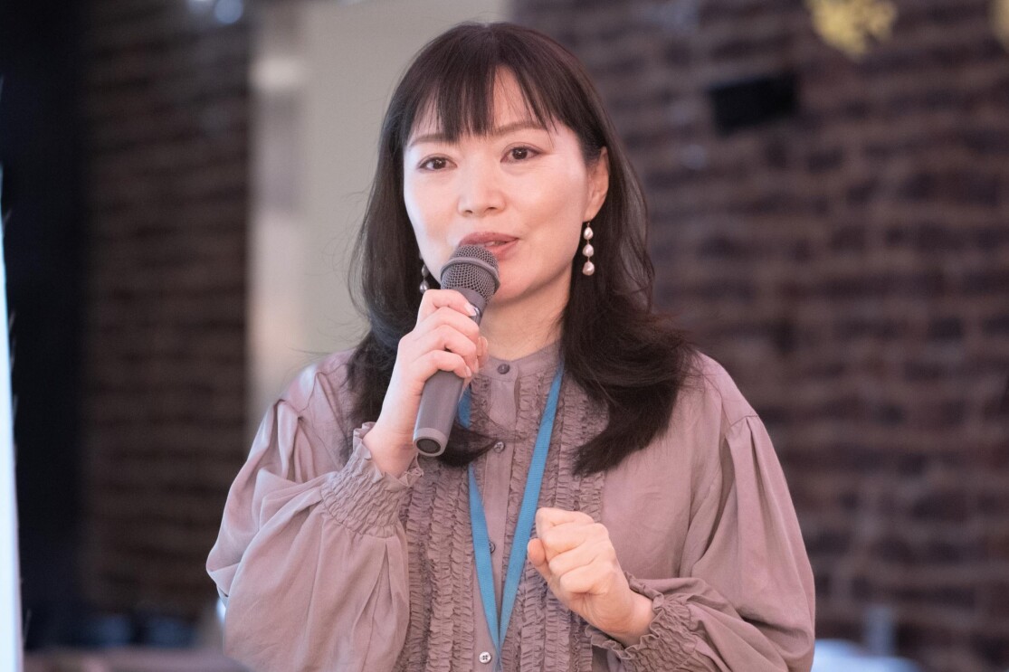 紫色のブラウスを着ている女性がマイクでお客さんに説明しているようだ。彼女はレンガの壁で囲まれた部屋にいる。