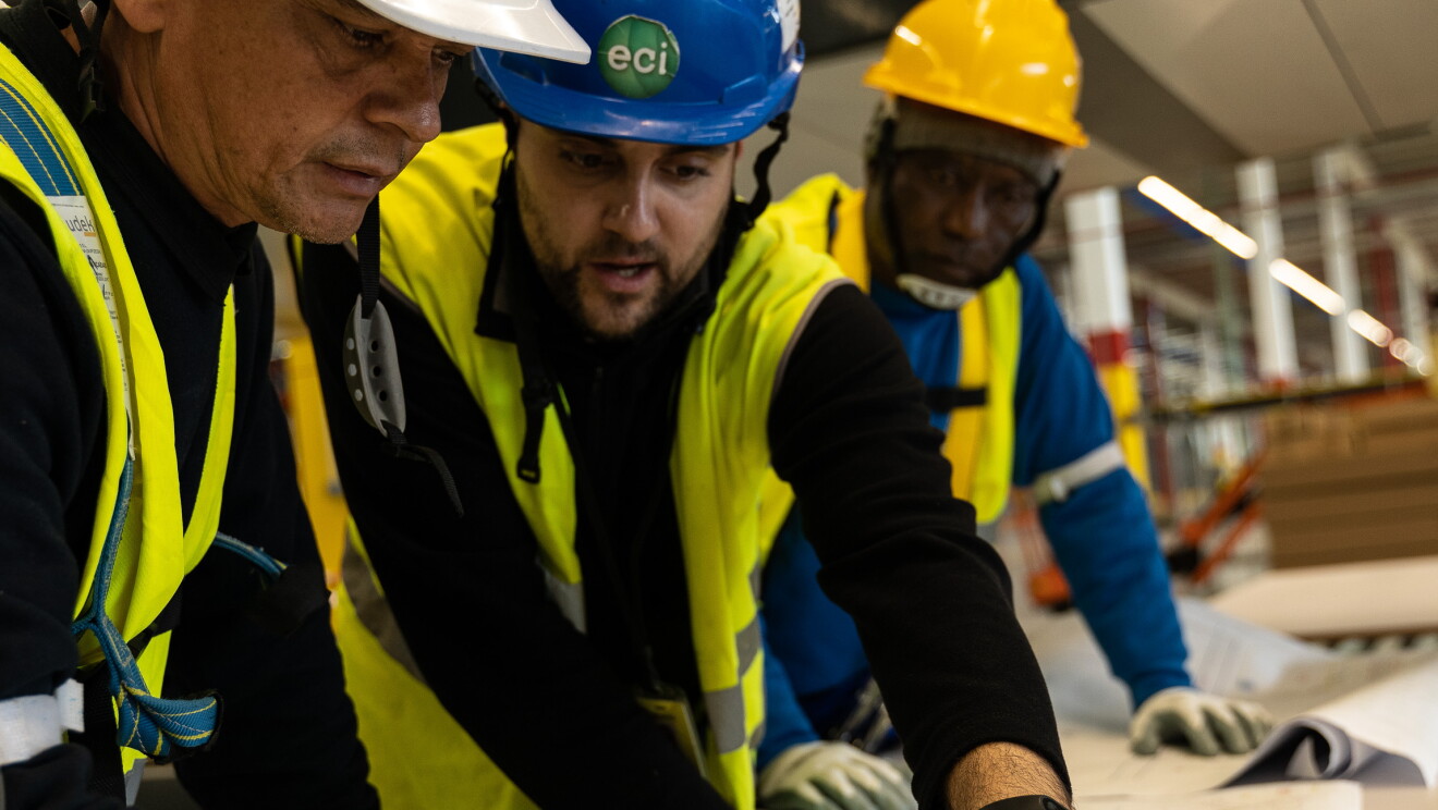Aparecen tres personas trabajando con un plano.  Las tres llevan cascos; blanco, azul y amarillo y chalecos amarillos y están trabajando en un plano que está sobre una mesa. Se ven cinco manos, tres de ellas con guantes blancos.