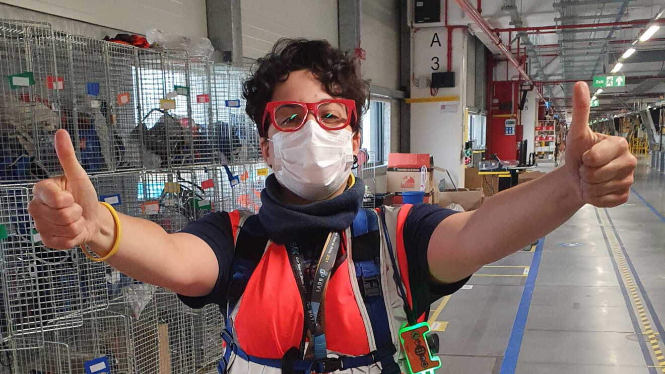 Mezzo busto di una dipendente Amazon in un magazzino. Ha le braccia alzate e con entrambe le mani fa il segno dell'OK con i pollici in alto. Ha i capelli corti e indossa un paio di occhiali da vista rossi. 