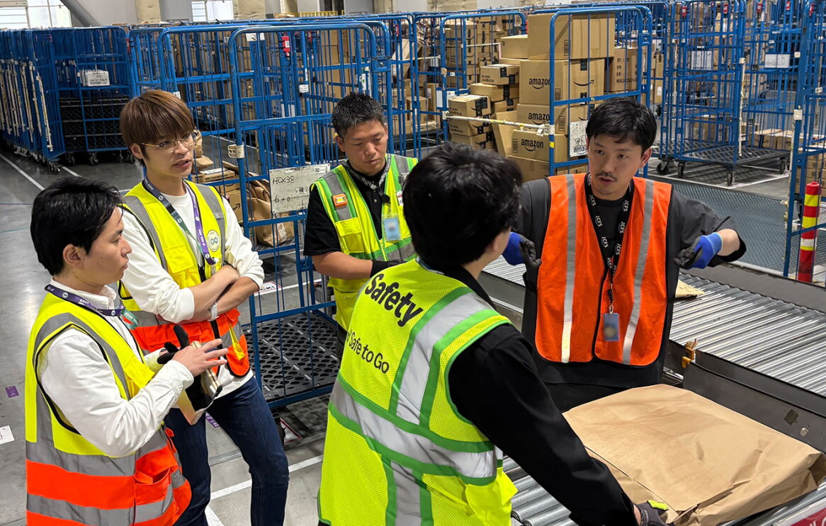 配送センターで、荷物を前に話し合う5人の人物。全員安全用の蛍光色ベストを着用している。背後に台車や荷物の山。