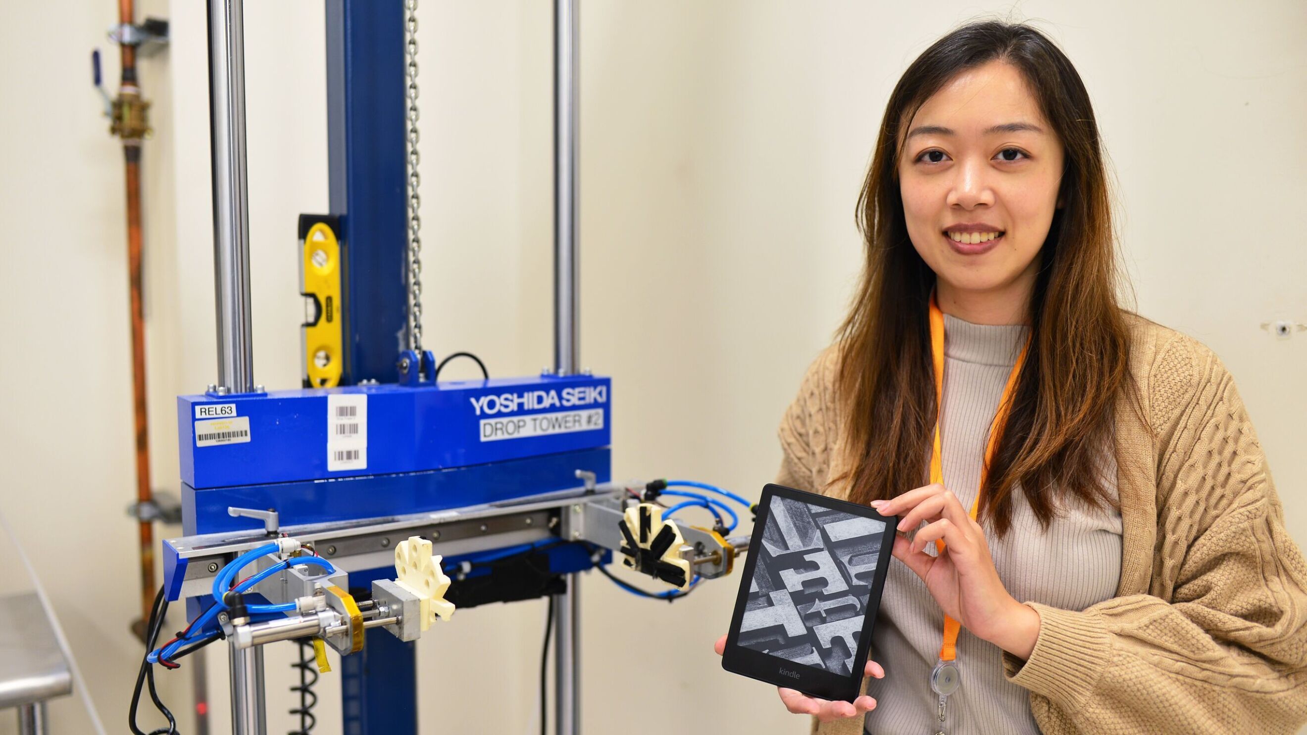 An image of a woman smiling in front of a drop test machine at the Amazon Lab126 Reliability Testing lab in California. She is holding a Kindle device.