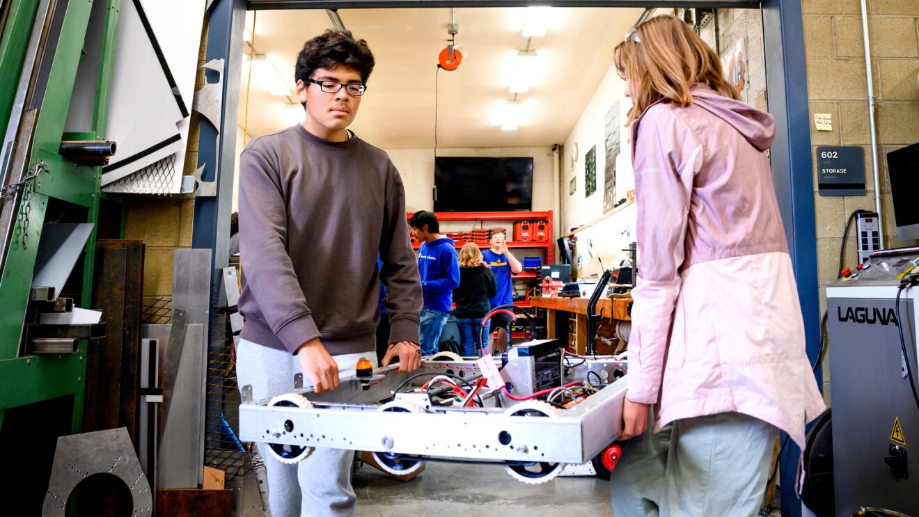 A photo of two students carrying a partially-built robot in a high school robotics lab.