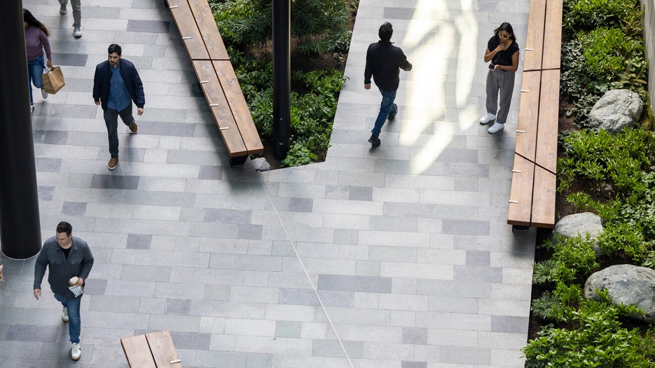 Four people are walking across a large sidewalk between Amazon buildings.