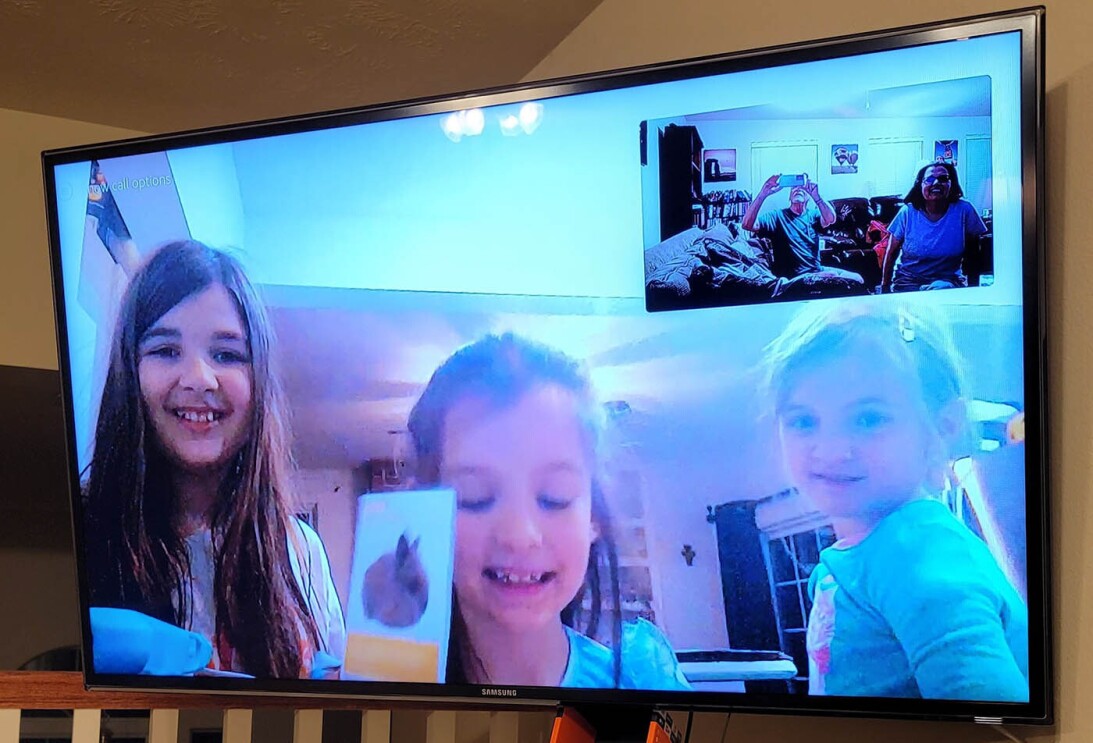 An image of a television screen with three children smiling on a video call with their family.