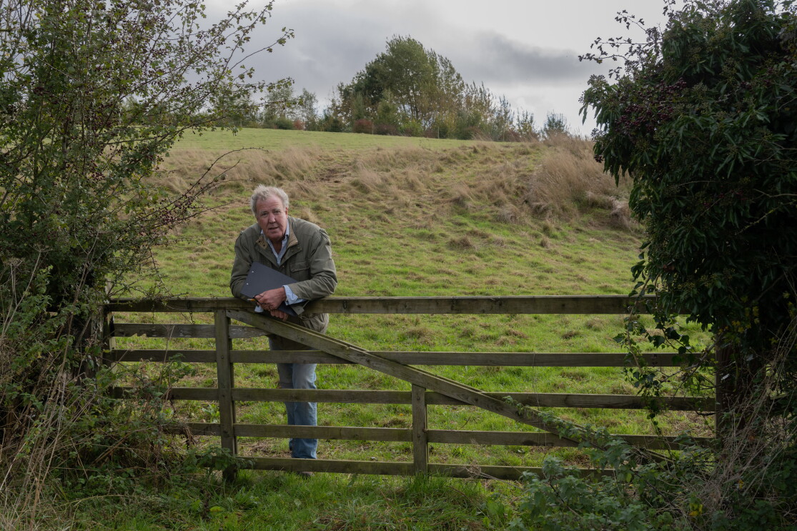 An elderly man holding a clipboard and leaning against a fence with a farm in the background 