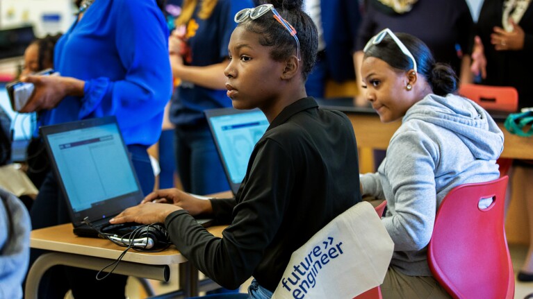 A photo of two students sitting at a desk in front of two laptops, listening to an instructor.