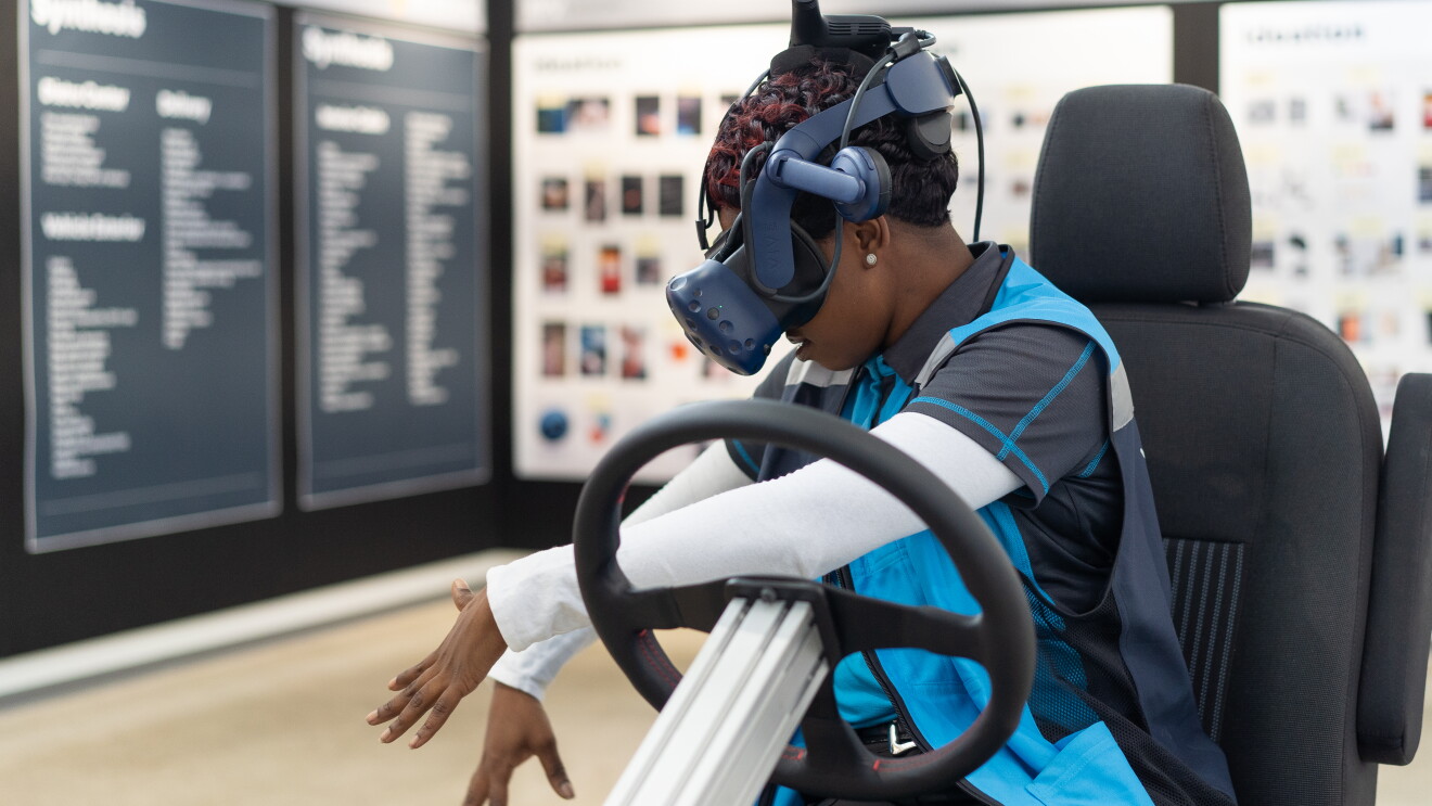 Donna indossa un visore per realtà virtuale mentre è seduta su una poltrona con un volante di fronte a sè. 