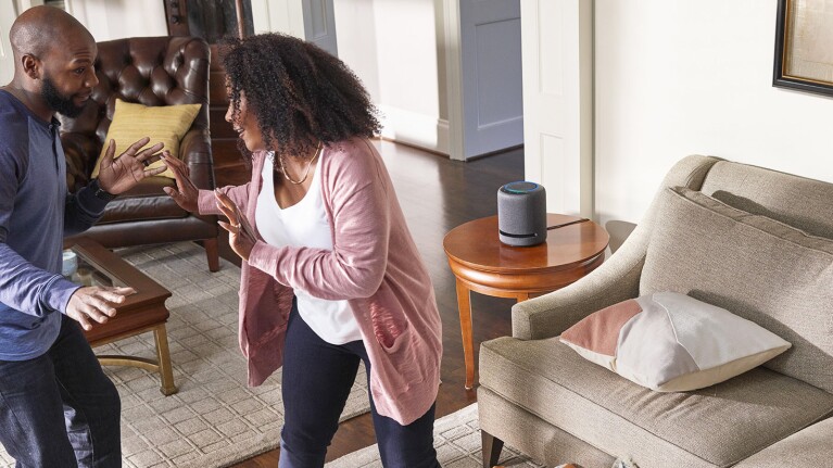 An image of a man and a woman dancing in a living room. There is an Amazon Echo device sitting on the side table next to a sofa.