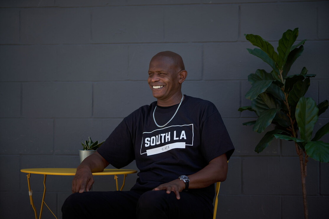 An image of a man smiling while sitting at a yellow outdoor table. His shirt says "South LA Cafe"
