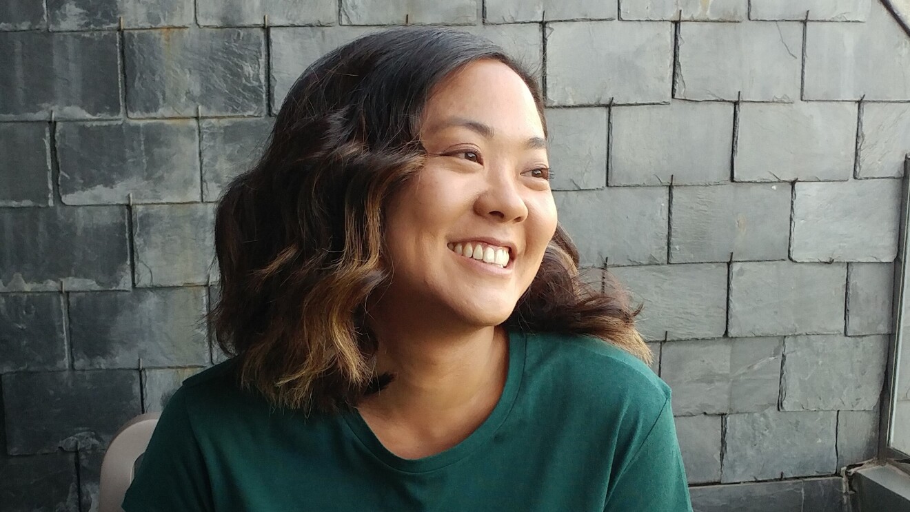 A photo of a woman smiling while looking away from the camera. She is standing in front of a brick wall wearing a green shirt.