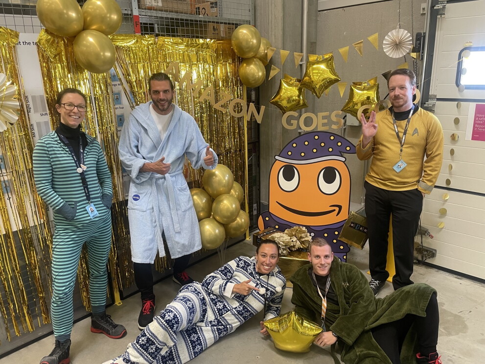 Fünf Mitarbeiter:innen vom Amazon Verteilzentrum Hamburg stehen in Pyjamas und Bademänteln vor dem „Amazon Goes Gold“ Banner.