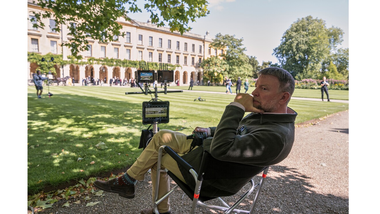 前庭に緑の芝生が広がる大きな建物。芝生の手前側の砂利道に置かれた監督椅子に座る男性。椅子の前には大小のモニターが3点設置されている。