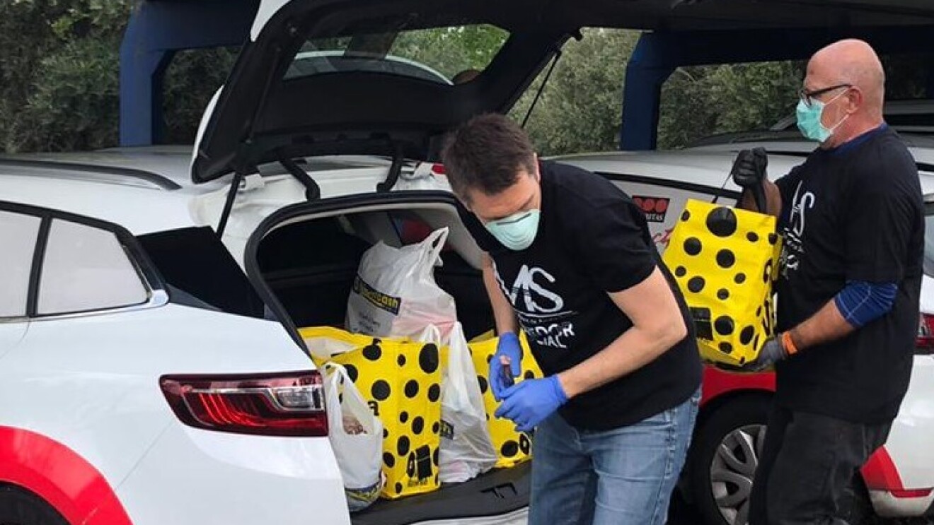 Manos de ayuda social reparte productos de primera necesidad gracias a la ayuda también de Securitas direct. Aparece en primer plano un coche de Securitas direct con dos de sus conductores que llevan una camiseta de Manos de ayuda socials de color negro. Los dos hombres de mediana edad llevan mascarilla y están colocando bolsas de color amarillo con lunares negro en el maletero de un coche. Están en un aparcamiento abierto.