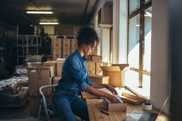 A woman sits on a step stool as she packages a product in a kraft box.