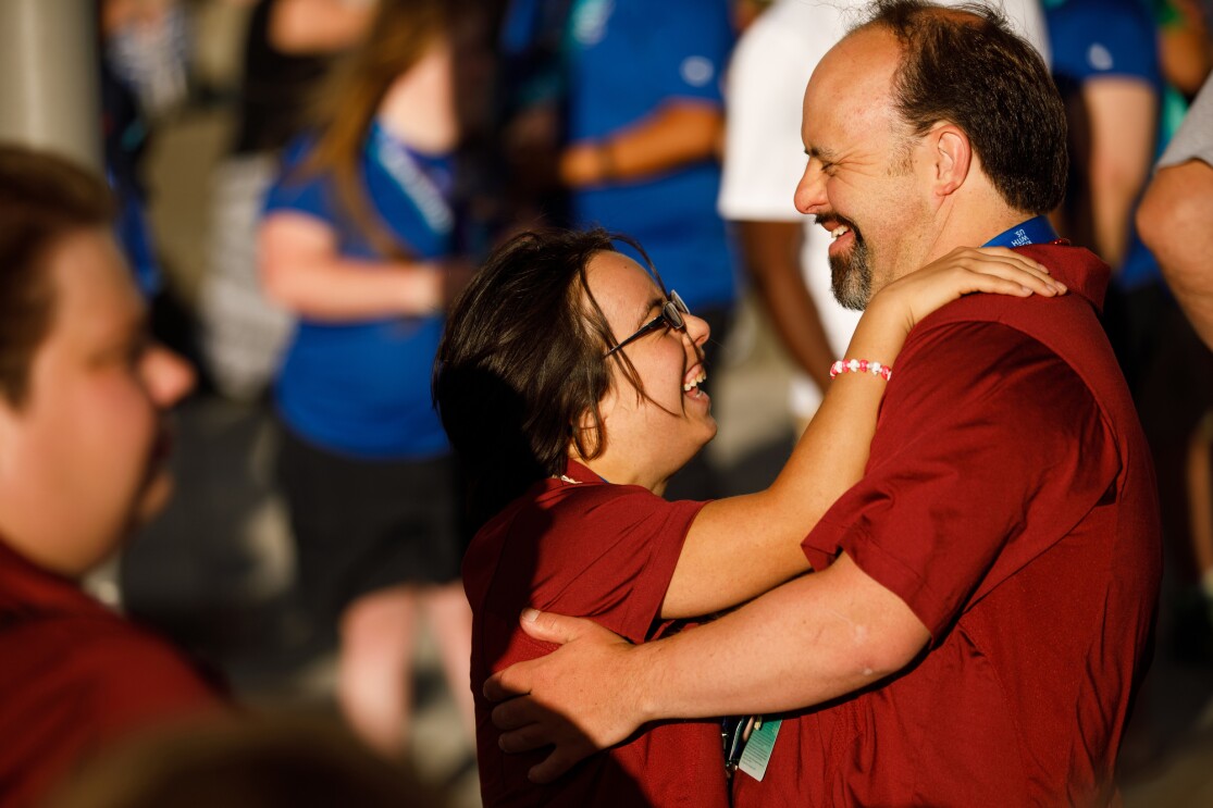 Two Special Olympics USA Games athletes (a male and female, both in red shirts) dance in the foreground, volunteers are seen standing in the background.