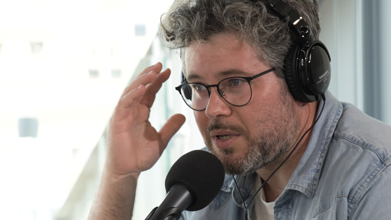 César Pascual, fundador de Cerveza La Virgen con una camisa  tejana azul y debajo una camiseta blanca de algodón con un dibujo que no se puede descifrar. Lleva gafas y tiene el pelo canozo de punto y una barba con canas. Sentado en una mesa delante de un micrófono y con unos auriculares puestos y una sonrisa. El brazo derecho alzado.  
