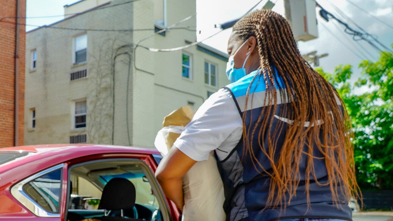 A delivery driver in an Amazon vest carries a bag of groceries to a car.