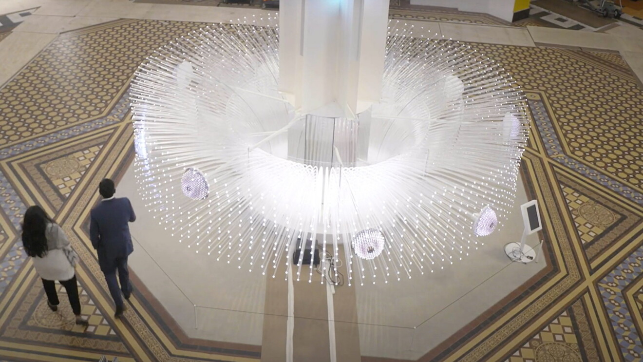An image of a large white structure with multiple glass shoots coming out of it. At the end of each shoot is a small light. There are small circles within the structure that have smaller clusters of lights with a purple tint. The structure stands in the middle of a museum, and two people are walking by it.