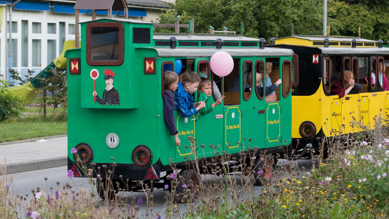 Kinder fahren in einer kleinen Lokomotive auf der Straße. Im Hintergrund ist ein Spielplatz, das Krankenhaus und eine Blumenwiese. Die Kinder schauen aus den Fenstern der Wagons und haben Spaß.