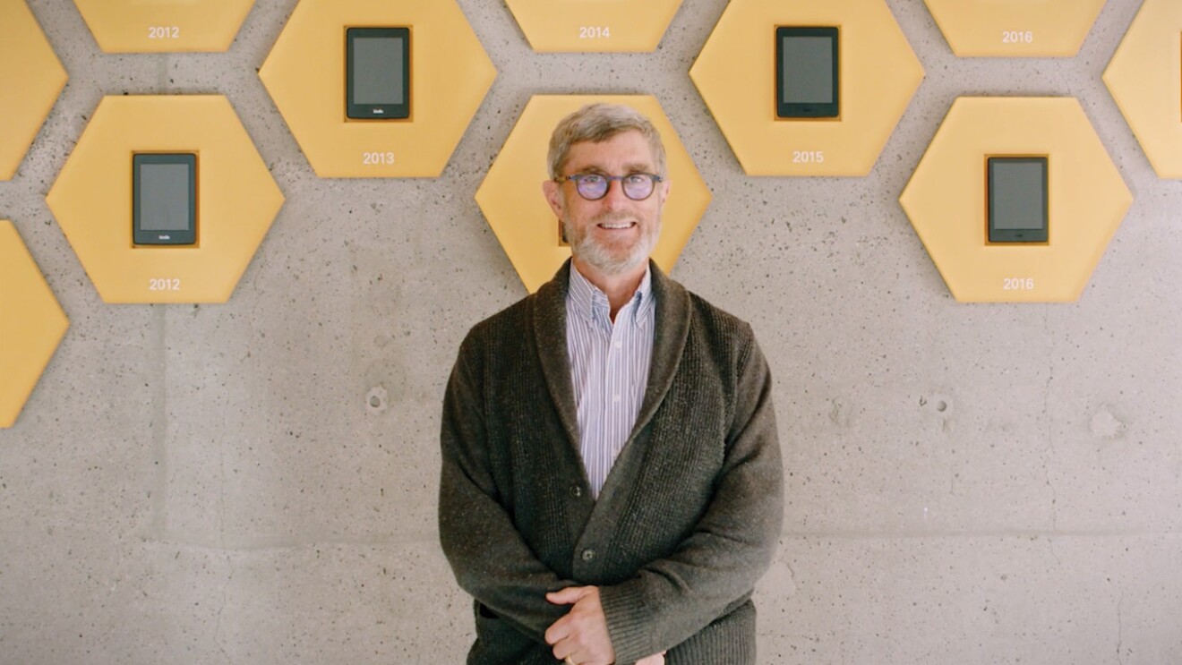 An image of Len Edgerly, host of The Kindle Chronicles, posing with a wall showing the different generations of Amazon's Kindle behind him. 