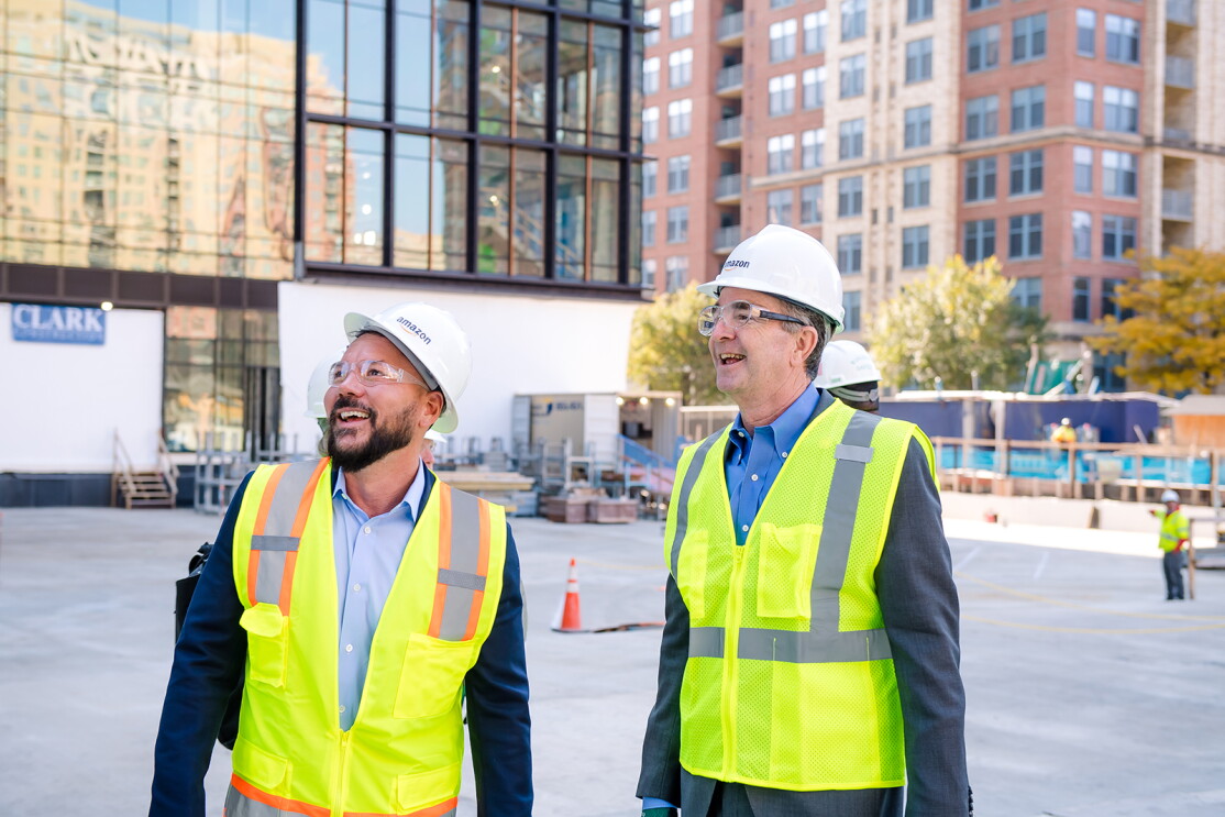 An image of people touring the construction site of Amazon's second headquarters while wearing hard hats that say "Amazon" and bright yellow safety vests.