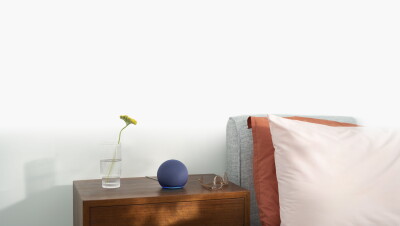 El Echo Dot, con la luz azul encendida, en una mesita de noche. Delante de una pared blanca, hay una mesita de noche de madera oscura. Encima hay un vaso con agua y una flor de color amarillo, el dispositivo Echo Dot y unas gafas. Al laso un cabecero de color gris, dos cojines con dos tonalidades de rosa y un cubrecamas de color verde.
