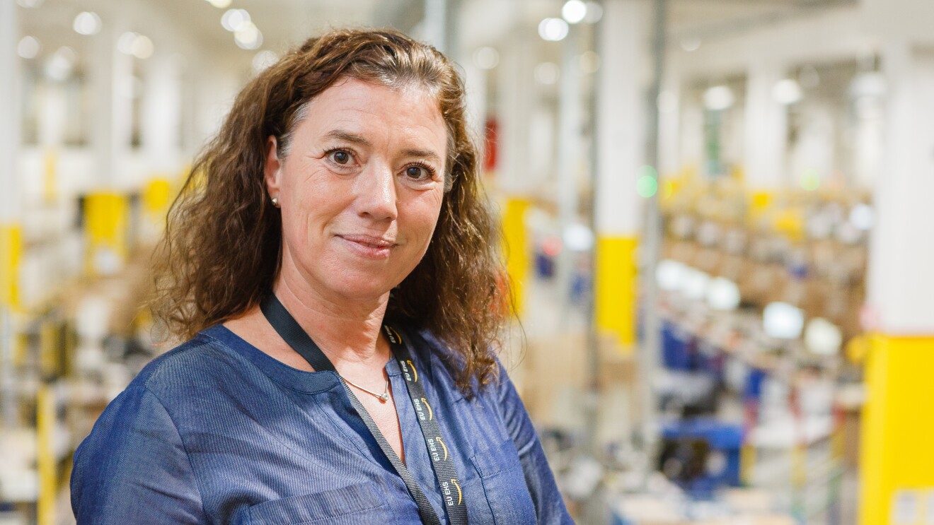 Eine braunhaarige Frau in blauer Bluse in einem Logistikzentrum (Aufnahme vor Pandemie).