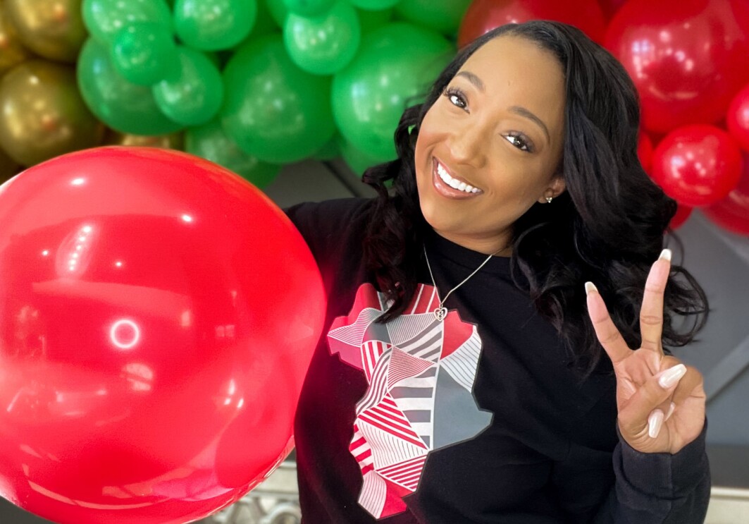 An image of a woman smiling for a photo while making a peace sign. She is standing in front of a balloon arch with green, red, black, and gold balloons, and holding a large red balloon under one arm.