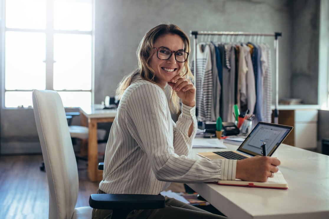 Una donna sorridente seduta nel suo ufficio mentre annota qualcosa su una agenda. Indossa degli occhiali da vista. Sullo sfondo, una rella con degli abiti appesi