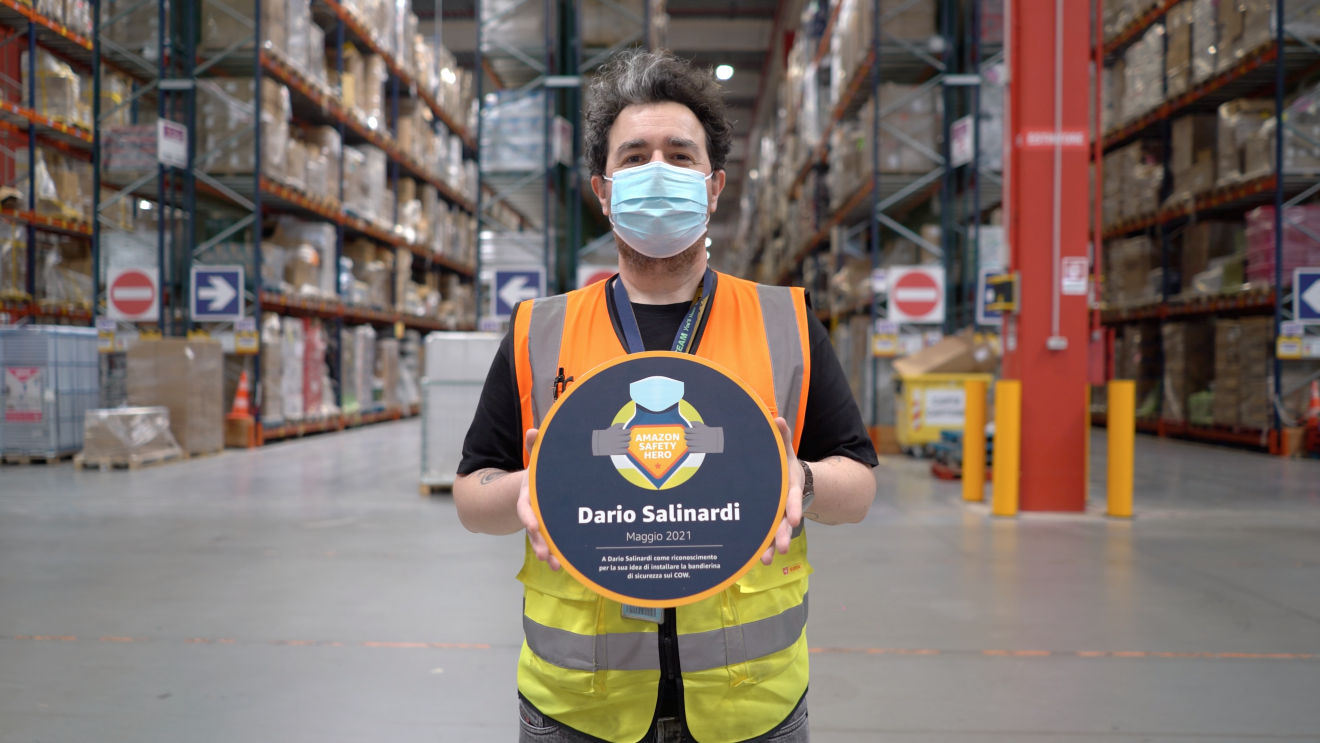 Uomo ripreso a mezzo busto in un magazzino Amazon. Indossa un giubbino catarinfrangente arancione e giallo, tiene tra le due mani e mostra alla camera un cartellone tondo con la scritta 'Dario Salinardi 2021' ed un logo che recita 'Amazon Safety Hero'. Indossa una mascherina chirurgica sul volto, ha i capelli neri con un ciuffo bianco. Alle sue spalle si intravedono gli scaffali di un magazzino Amazon.