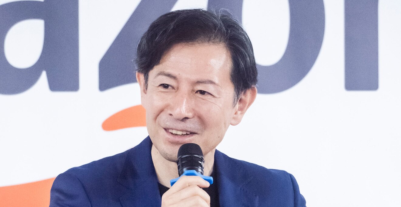 大きなアルファベットが書かれた壁を背に、マイクを持って話をする男性。ネイビーのスーツを着用、丸襟のインナー。