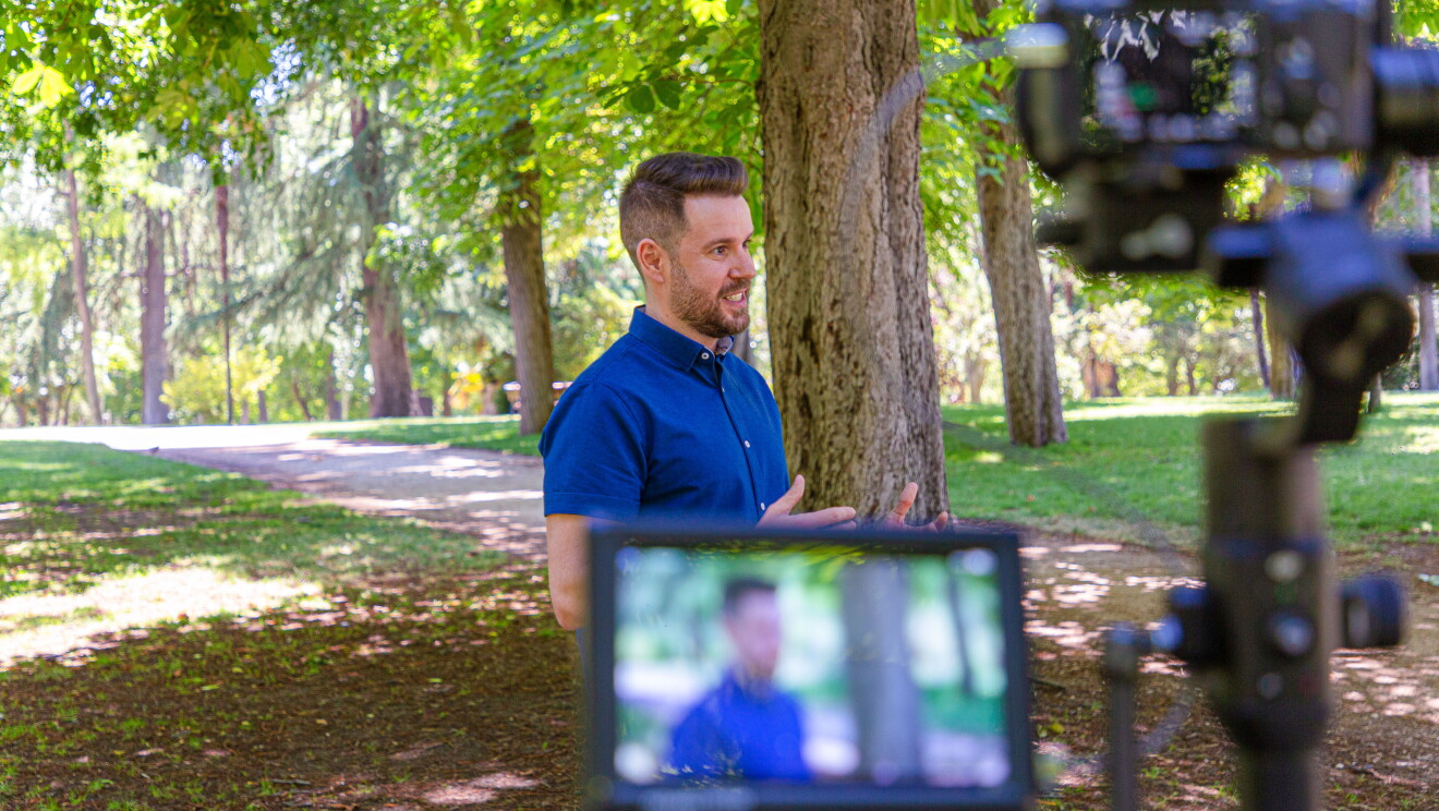Paul Pen en un momento de la grabación de la historia en el Parque de El Retiro (Madrid). En primer plano sale la cámara con la pantalla y el protagonista desenfocado. En segundo plano pero enfocado está Paul Pen hablando a cámara. Lleva una camisa azul y unos pantalones tejanos. De fondo se ven los árboles del parque. 