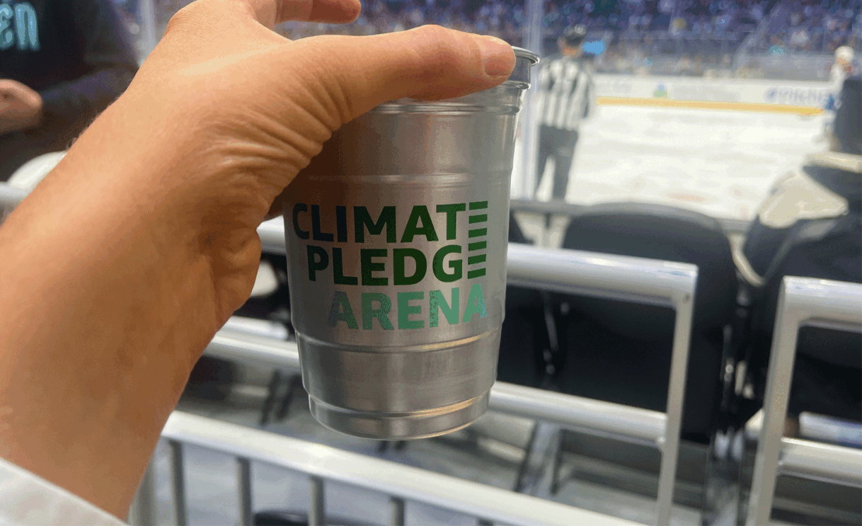 An image of a hand holding silver cup that says "Climate Pledge Arena" on it. In the background is an NHL ice rink.