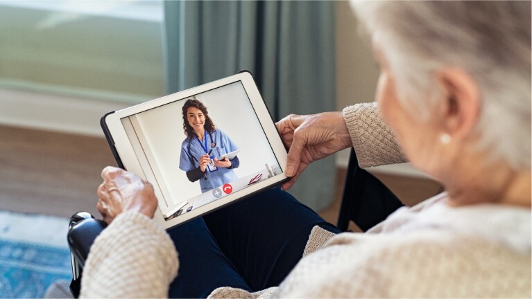 elderly person looking down at a tablet screen which shows a video of a clinician consulting during a health appointment