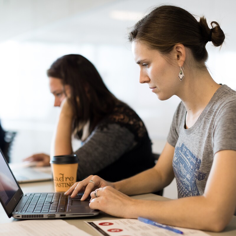 A woman wearing a t-shirt works on a laptop computer. Behind her, other individuals seated at work stations also work on laptop computers. Images were captured pre-COVID-19.