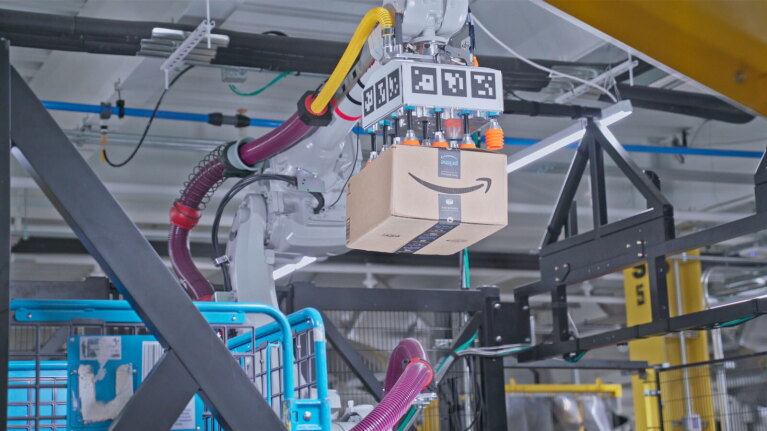 An image of a gray robotic arm with purple and yellow tubes on it. It is lifting an Amazon box which it will place in a blue crate below it.