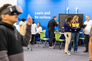 A photo of students visiting the AWS Think Big Space in at J. Lupton Simpson Middle School in Leesburg, Virginia. There is a student in the forefront of the photo trying on virtual reality goggles, and students talking with several teachers in the background.