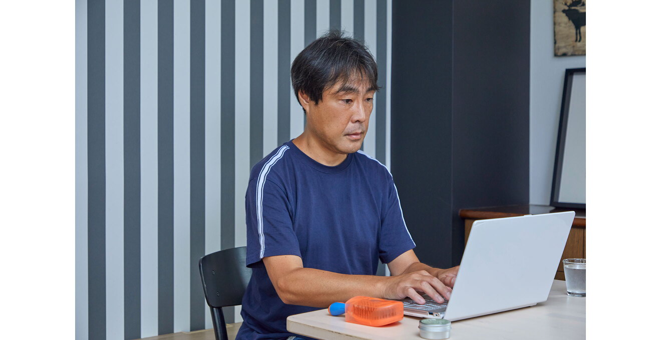 デスクに着席してノートパソコンで作業する男性。背後の壁は明暗差のつよいストライプ。