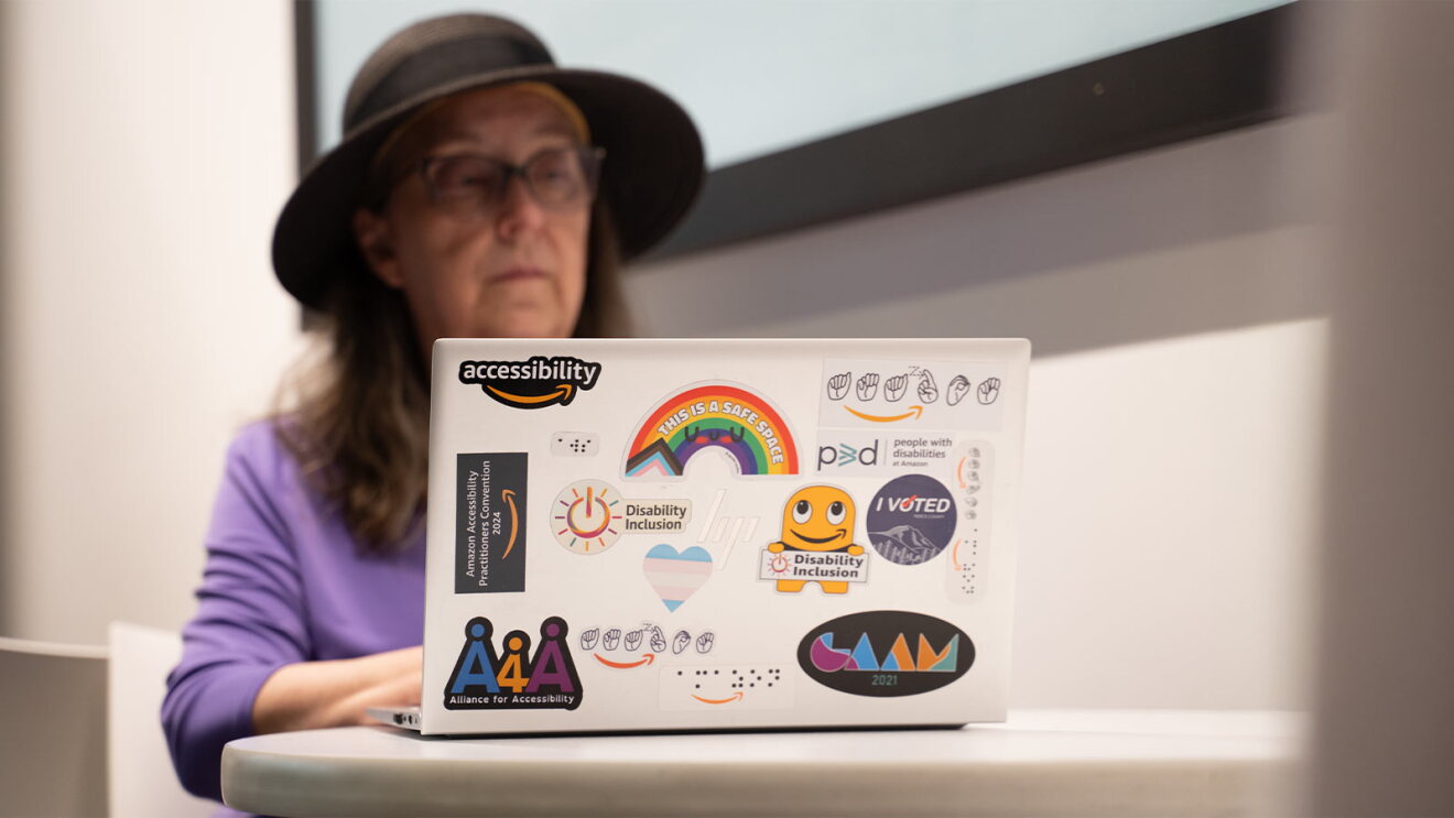 Laptop covered in accessibility and disability inclusion stickers, foreground of person in purple shirt and hat