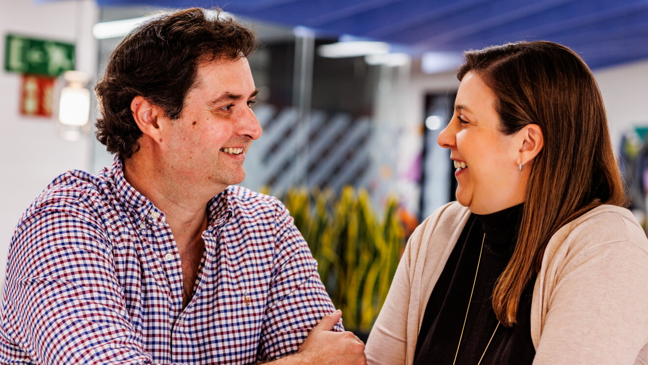 Ane y Pablo posando en las oficinas corporativas en Madrid para la sesión de fotos del post que tenemos entre manos. Los dos están sentados, con los brazos cruzados y sonriendo, Apoyan las manos en la mesa donde tienen dos vasos de café. Él lleva una camisa de cuadros de color rojo y azul y ella lleva el pelo suelto, un jersey de cuello alto de color negro y por encima una chaqueta de punto. De fondo se ven unas plantas y unos cristales de una oficina.