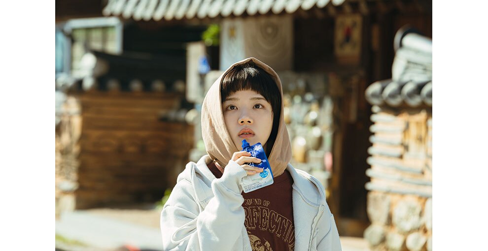 明るい屋外に立ち、手にドリンクのパウチを持った人物。パーカーのフードを着用している。背後には韓国の伝統的な建物の外観。