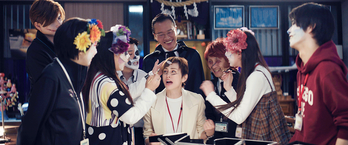 神棚があるオフィスのような室内、骸骨のメイクや仮装をした9人の人物。中央の女性だけ座っていて、左側に立った女性が人物がメイクを施している。