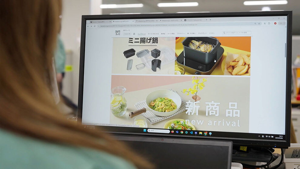 モニターに映された商品販売サイトの画面。ミニ揚げ鍋の商品画像とその下に「新商品」の文字が表示されている。左手前にモニターを見る人物の後ろ姿があるが、ピントは画面に合っている。