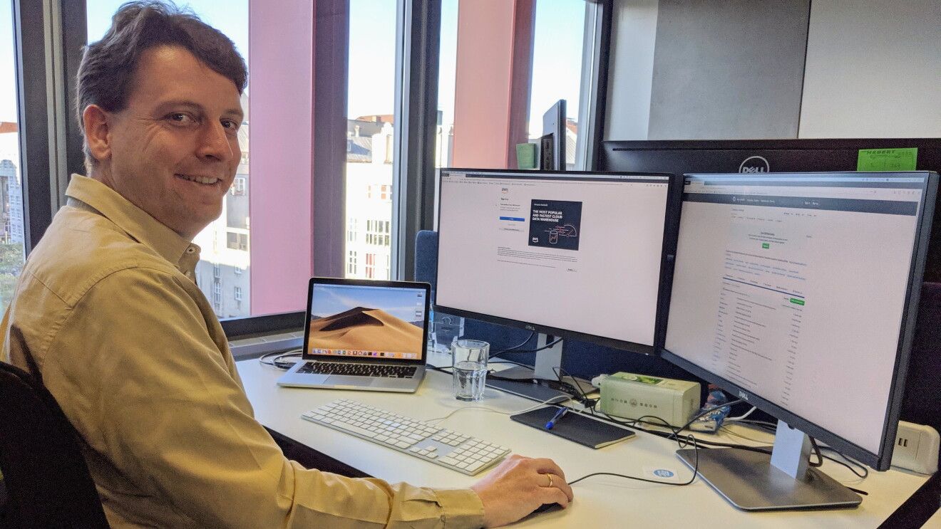 Ein Mann mit kurzer braunen Jahren sitzt an einem Schreibtisch mit Apple Laptop und 2 Monitoren.