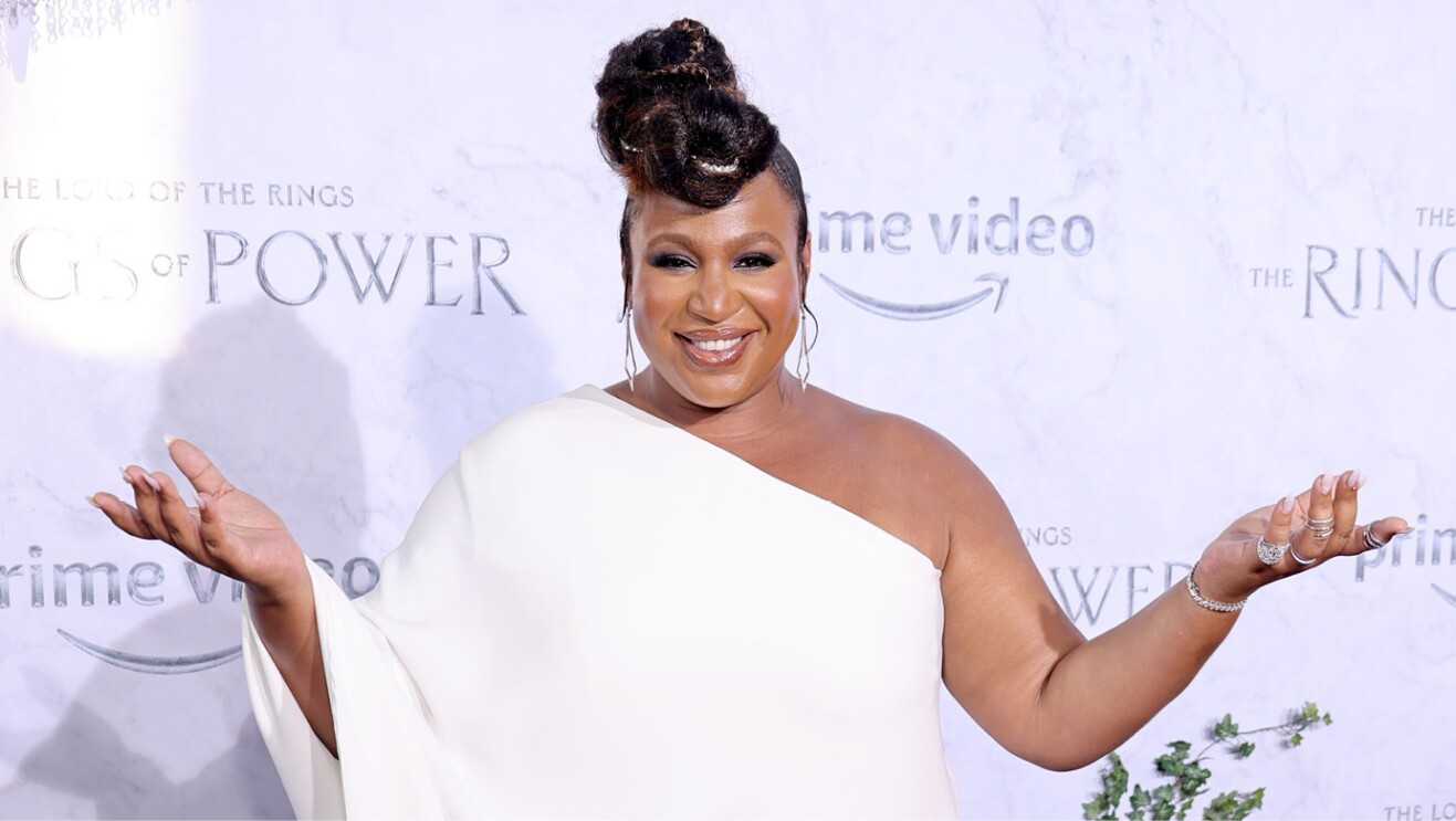 A woman smiles for a photo holding her arms out. The Lord of the Rings branding is behind her on a backdrop of the red carpet.
