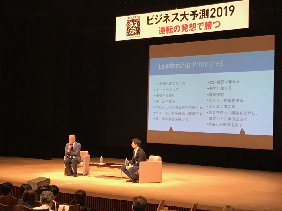 薄暗いホールのステージ上でソファに座って話をする2人の人物。ステージ奥のスクリーンにスライドが表示されている。